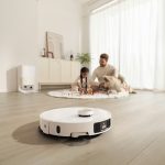 Saugroboter fährt über hellen Holzboden, im Hintergrund die Reinigungsstation und eine Familie im Wohnzimmer