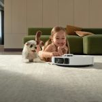 Saugroboter auf einem Teppich im Wohnzimmer, daneben ein Kind mit Hund