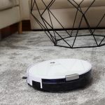 Weißer Saugroboter auf einem grauen Hochflorteppich vor einem Sofa